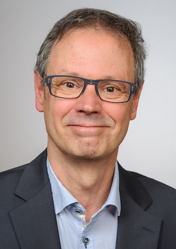 Porträtfoto Bernhard Kiesel: blickt lächelnd in die Kamera, kurze graue Haare, anthrazitfarbene Brille, hellblaues Hemd, anthrazitfarbenes Jacket