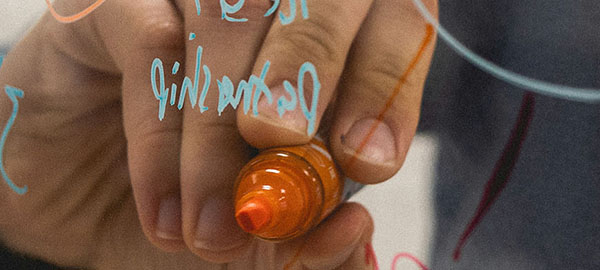 Eine Hand schreibt mit orangefarbenen Edding von hinten auf eine Glasscheibe, auf der bereits einzelne Wörter in Türkis stehen.