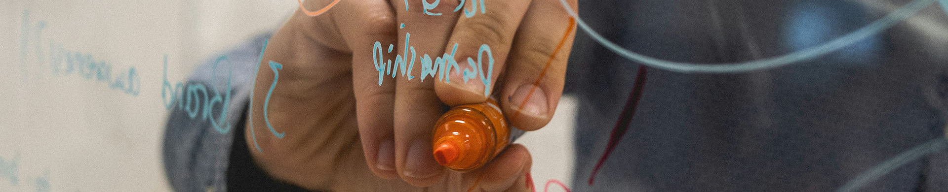 Eine Hand schreibt mit orangefarbenen Edding von hinten auf eine Glasscheibe, auf der bereits einzelne Wörter in Türkis stehen.