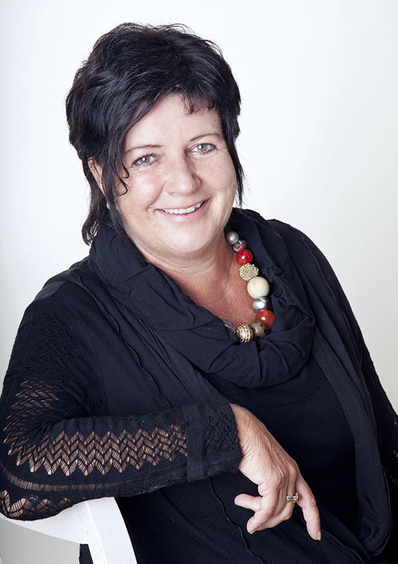 Porträtfoto Doris Ebner-Fiedler: sitzt seitlich auf einem Stuhl, schwarze kurze Haare, schwarzes Oberteil, Kette mit großen Kugeln