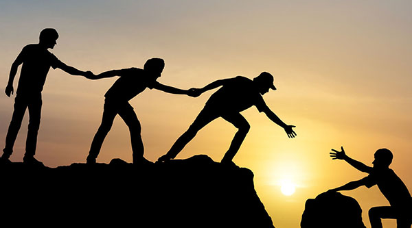 Die Silhouette zeigt drei Personen auf einem Felsen, die sich an den Händen halten und einer vierten Person die Hand reichen, die einen kleineren Felsen hochklettert